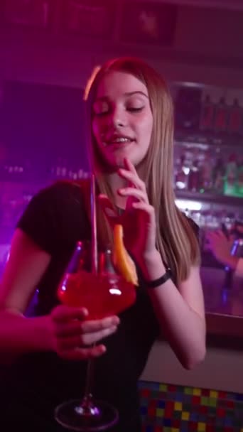 Bella ragazza balla in un nightclub con un cocktail rosso tra le mani. Sullo sfondo c'è il bancone del bar e il barista prepara bevande alcoliche usando il fuoco. Rallentatore, Video verticale - Filmati, video