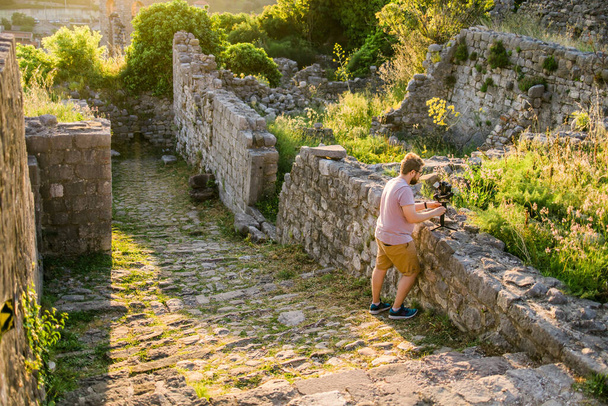 Cameraman em roupa de verão com gimbal e câmera de cinema em suas mãos fazendo vídeo de viagem com a velha natureza dos Balcãs da Europa em segundo plano. - Foto, Imagem