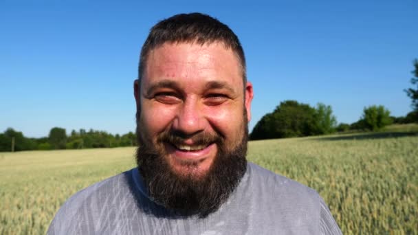 Portrait of agronomist with beard against background of green wheat. Happy smiling farmer looks into camera standing at barley meadow. Concept of agriculture and agronomy business. Close up. - Footage, Video