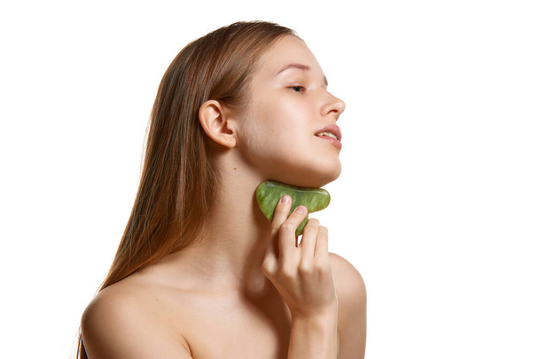 Portrait of young woman doing neck massage with natural stone scraper to tighten oval of face and remove double chin against white background. Skin care treatment. Concept youth, natural beauty. Ad - Photo, Image