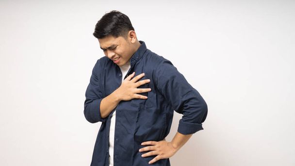 Fiatal ázsiai férfi megérintette a mellkasát fájdalmas arckifejezéssel. Szívroham gesztus fehér háttér - Fotó, kép