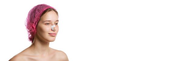 Een spandoek. Portret van een aantrekkelijke vrouw in roze pet met pleisters op de neus na neuscorrectie tegen witte studio achtergrond met kopieerruimte. Concept van de geneeskunde, schoonheid injecties, lichaam plastische chirurgie. - Foto, afbeelding