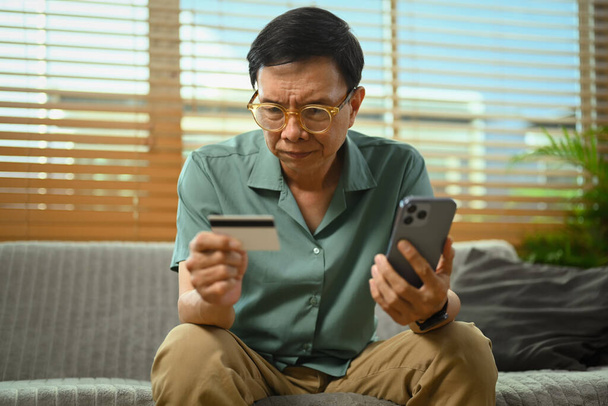 Homme âgé sérieux détenant une carte de crédit et un téléphone mobile, effectuant des transactions en ligne - Photo, image