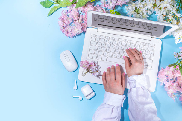 春のオフィスの職場、ブログフラットレイアウトの背景。白いノートパソコン、ヘッドフォン、タブレット、春の花の花束ライトブルーの背景、女の子の手はノートパソコンに入力して. - 写真・画像