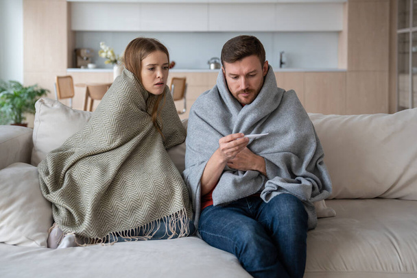 Sairas pari istuu sohvalla kääritty huopa, tarkistaa lämpötila lämpömittari. Vaimo ja aviomies, joilla on hengitystieinfektion, influenssan, kuumeen oireita ja jotka tarvitsevat hoitoa ja eristäytymistä - Valokuva, kuva