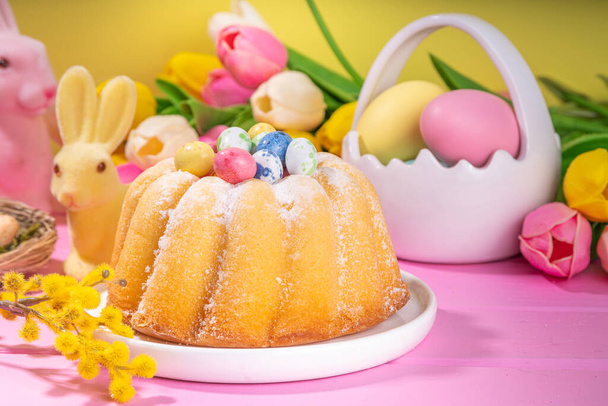 Gâteau rond traditionnel de Pâques. Cuisson de Pâques traditionnelle sucrée avec des saupoudres de sucre, oeufs de Pâques colorés, fleurs de printemps, sur fond jaune rose, espace de copie coloré - Photo, image