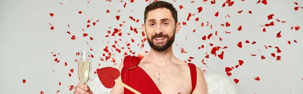 cheerful bearded man dressed as cupid with arrow and champagne under red confetti on grey, banner - Photo, Image