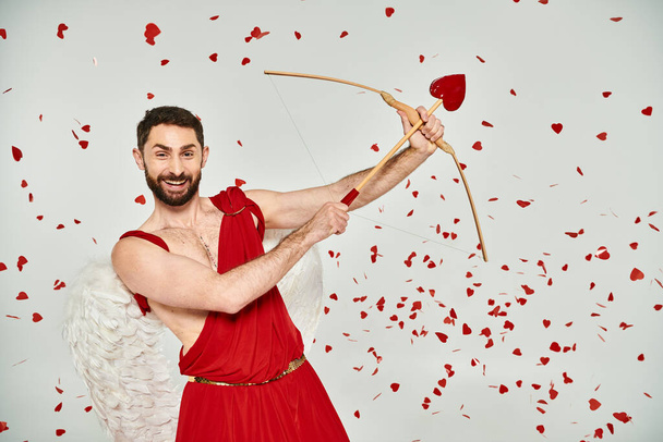 excitado barbudo hombre Cupido arqueo bajo confeti en forma de corazón en gris, San Valentín fiesta - Foto, imagen