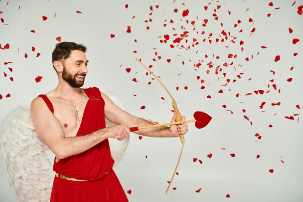 Улыбающийся бородатый амур стреляет из лука стрелой в форме сердца под красными конфетти, День Святого Валентина - Фото, изображение