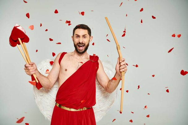 bearded man in cupid costume with heart-shaped arrows and bow screaming from excitement on grey - Photo, Image