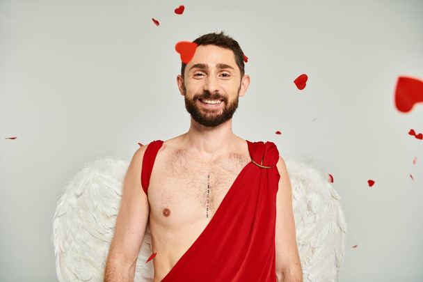 hombre barbudo en traje de Cupido y alas sonriendo a la cámara bajo confeti rojo en forma de corazón en gris - Foto, imagen