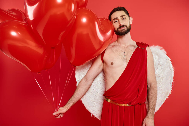 hombre de Cupido soñador y feliz con globos en forma de corazón mirando a la cámara en rojo, San Valentín día - Foto, imagen
