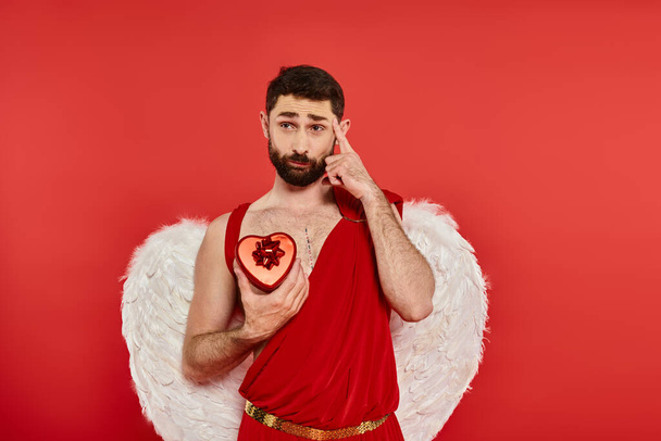 přemýšlivý vousatý muž v amorčím kostýmu s dárkovou krabicí ve tvaru srdce ukazující na jeho hlavu na červenou - Fotografie, Obrázek