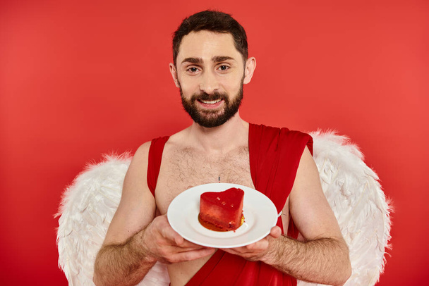 szczęśliwy brodaty mężczyzna w kostium Kupidyna gospodarstwa pyszne ciasto w kształcie serca i patrząc na aparat fotograficzny na czerwono - Zdjęcie, obraz