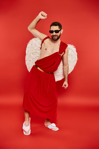 Aşk tanrısı kostümü giymiş heyecanlı bir adam ve kırmızı Sevgililer Günü partisinde dans eden güneş gözlüğü. - Fotoğraf, Görsel