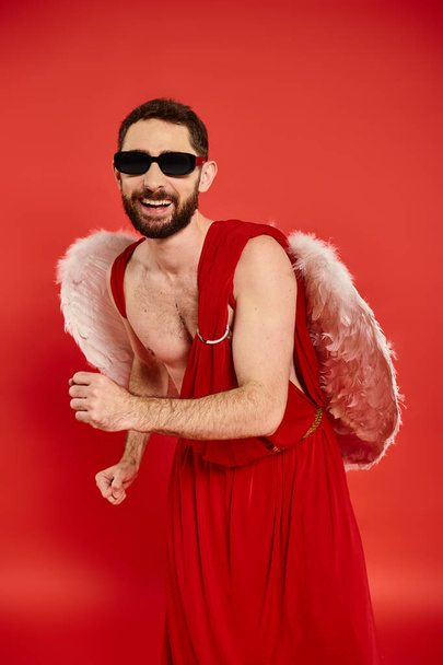 homem barbudo peculiar em óculos de sol e traje de cupido dançando no vermelho, festa temático dia de São Valentim - Foto, Imagem