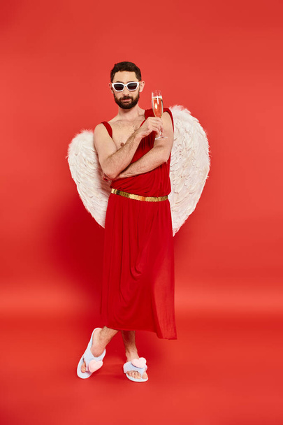 chladný vousatý muž v Amorském kostýmu pózující se sklenkou šampaňského na červeném, Svatý Valentýn - Fotografie, Obrázek
