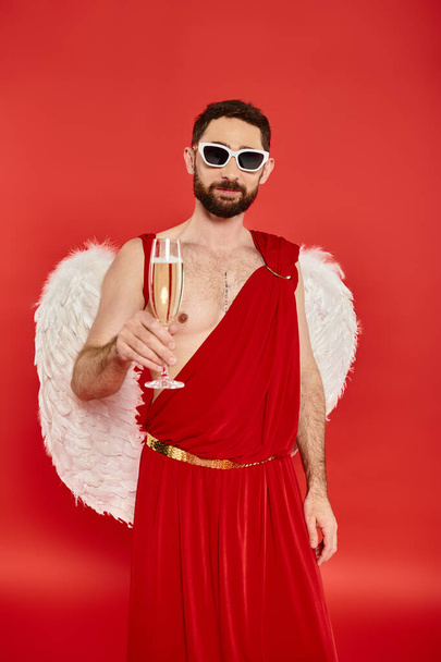 man met baard in zonnebril en cupido kostuum toosten met champagne op rood, Saint Valentines dag - Foto, afbeelding