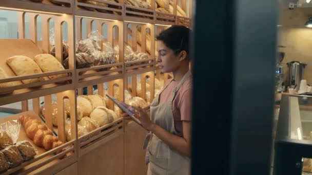 Nuori naispuolinen työntekijä esiliinassa katselee leipää esillä hyllyillä ja käyttää digitaalista tablettia tehdessään inventaariota leipomossa - Materiaali, video