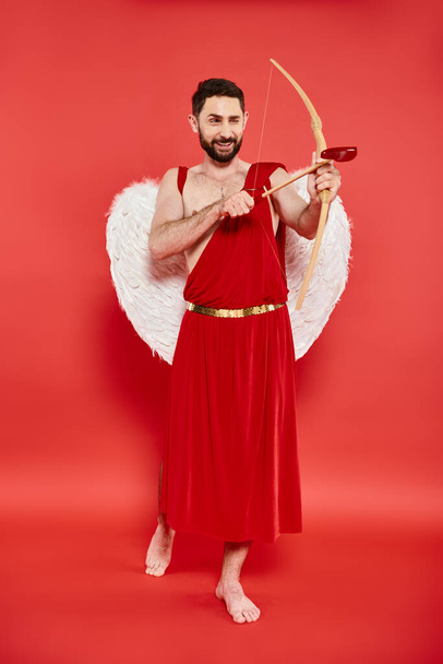 full length of smiling barefoot man in cupid costume looking away and archering on red backdrop - Photo, Image