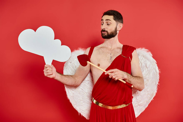 pensive cupid man with heart-shaped arrow looking at white empty thought-bubble on red backdrop - Photo, Image
