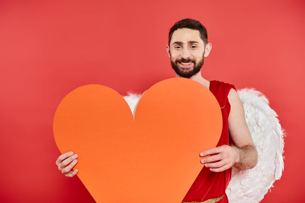 joyful bearded man with cupid wings holding huge orange heart on red, Saint Valentines day - Photo, Image