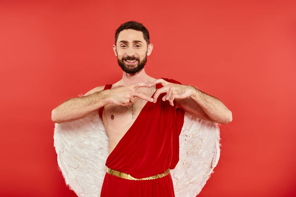 pleased bearded cupid man showing heart sign with fingers and smiling at camera on red backdrop - Photo, Image