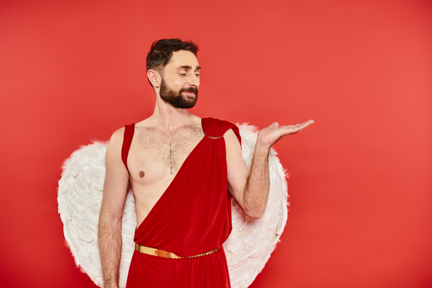 sonriente barbudo hombre Cupido señalando con la mano con la palma abierta en rojo, San Valentín fiesta de disfraces - Foto, imagen