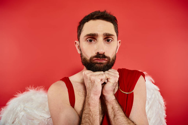 portret van gefrustreerde bebaarde man verkleed als cupido kijkend naar camera op rood, st valentines concept - Foto, afbeelding