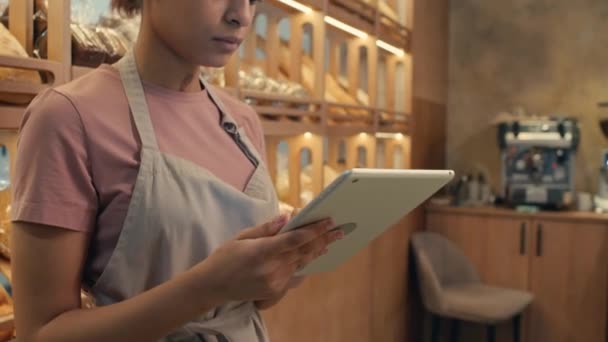 Plan incliné de la jeune femme dans le tablier en utilisant une tablette numérique et en regardant les marchandises sur les étagères d'exposition tout en faisant l'inventaire dans la boulangerie - Séquence, vidéo