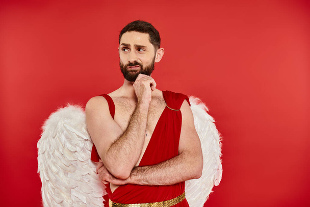 hombre barbudo disgustado en traje de Cupido cogido de la mano cerca de la barbilla y mirando hacia otro lado en el fondo rojo - Foto, imagen