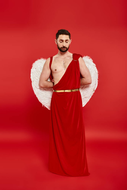in voller Länge von nachdenklichen bärtigen Mann in Amor-Kostüm stehend mit den Händen hinter dem Rücken auf rot - Foto, Bild