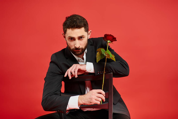 Selbstbewusster Mann im schwarzen Anzug sitzt auf Stuhl mit Rose und blickt am Valentinstag in die Kamera - Foto, Bild