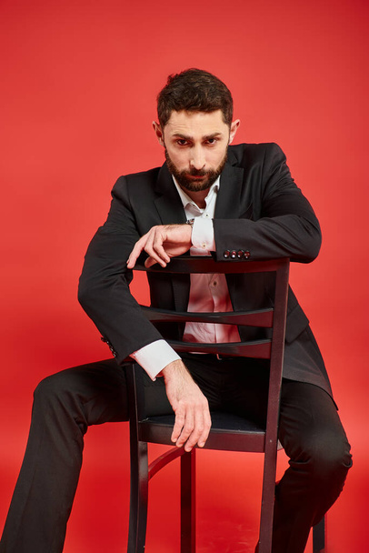 σοβαρός κομψός άνδρας κάθεται στην καρέκλα και κοιτάζει την κάμερα σε κόκκινο, επιτυχημένος επιχειρηματίας - Φωτογραφία, εικόνα