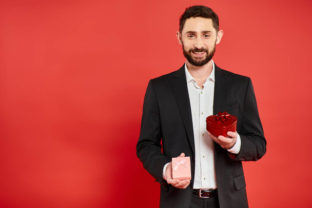 alegre hombre de negocios barbudo en traje elegante negro sosteniendo cajas de regalo en rojo, regalos de San Valentín - Foto, imagen