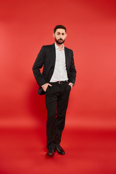 confident and stylish businessman in black suit standing with hands in pockets on red, full length - Photo, Image