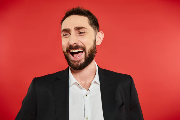 portrait of excited bearded businessman in black suit looking away and laughing on red backdrop - Photo, Image