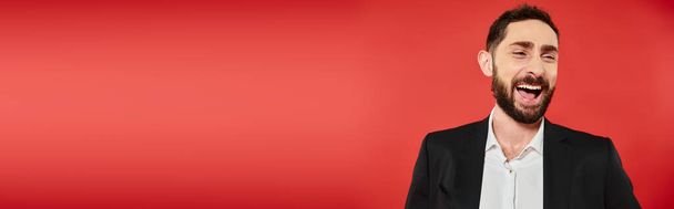 retrato de homem barbudo alegre em terno elegante preto olhando para longe e rindo no vermelho, banner - Foto, Imagem