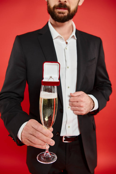 cropped view of man in black suit with champagne glass and gift box with jewelry ring on red - Photo, Image