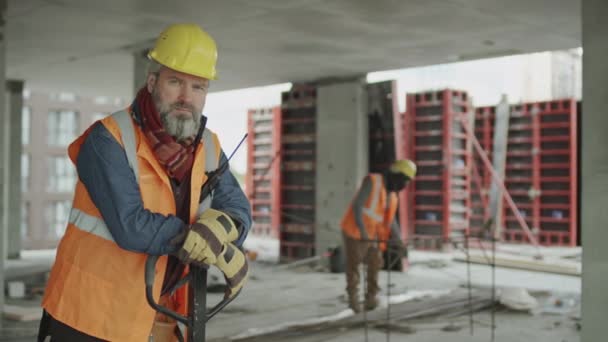Портрет среднего медлительного роста кавказского строителя среднего возраста, смотрящего в камеру, опираясь на тележку с поддонами на бетонной стройке - Кадры, видео