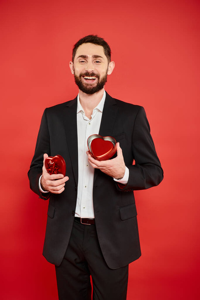 excited bearded man in black suit with heart-shaped gift box laughing on red, st valentines day - Photo, Image