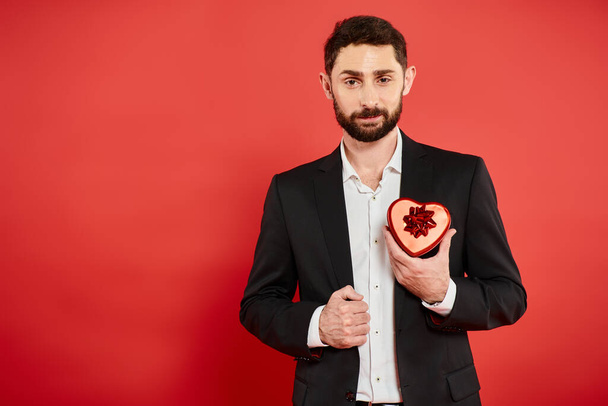 soddisfatto uomo d'affari barbuto in abito nero con scatola regalo a forma di cuore su rosso, San Valentino - Foto, immagini
