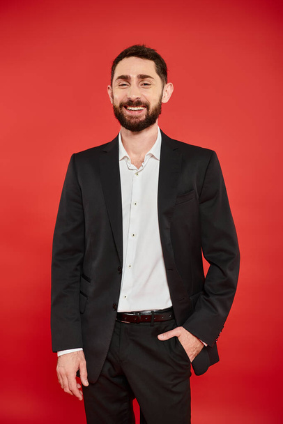 cheerful bearded businessman in black suit posing with hand in pocket and laughing on red backdrop - Photo, Image