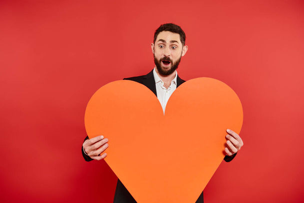 Şaşırmış sakallı adam, ağzı açık ve turuncu kağıt kalpli kırmızı, Aziz Sevgililer Günü konsepti - Fotoğraf, Görsel