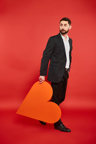 elegant man in black suit walking with huge paper heart and looking back on red, st valentines day - Photo, Image