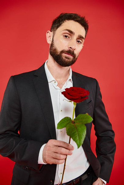 magabiztos jóképű férfi fekete öltöny piros rózsa néz kamera, Szent Valentin nap koncepció - Fotó, kép
