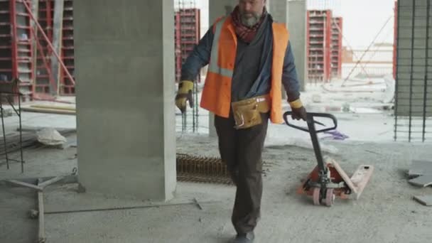 Powolniak dojrzałego białego robotnika budowlanego w twardej czapce i kamizelce z ręcznym wózkiem paletowym wzdłuż niedokończonego betonowego placu budowy - Materiał filmowy, wideo