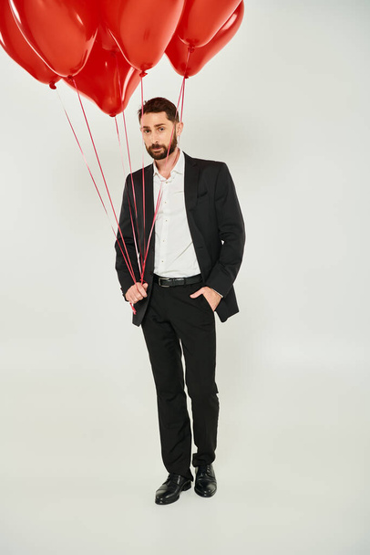 handsome bearded man in black suit with red heart-shaped balloons and hand in pocket on grey - Photo, Image