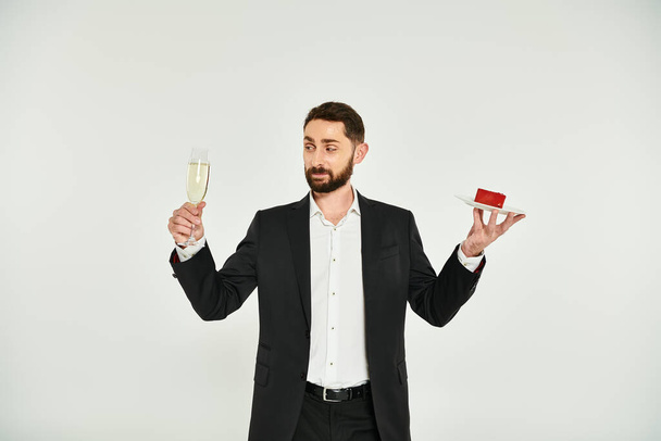 stylowy mężczyzna w czarnym garniturze z kieliszkiem szampana i tortem w kształcie serca na szarym, Walentynki - Zdjęcie, obraz