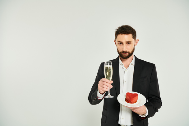 bel homme élégant avec verre de champagne et délicieux gâteau en forme de coeur regardant la caméra sur gris - Photo, image